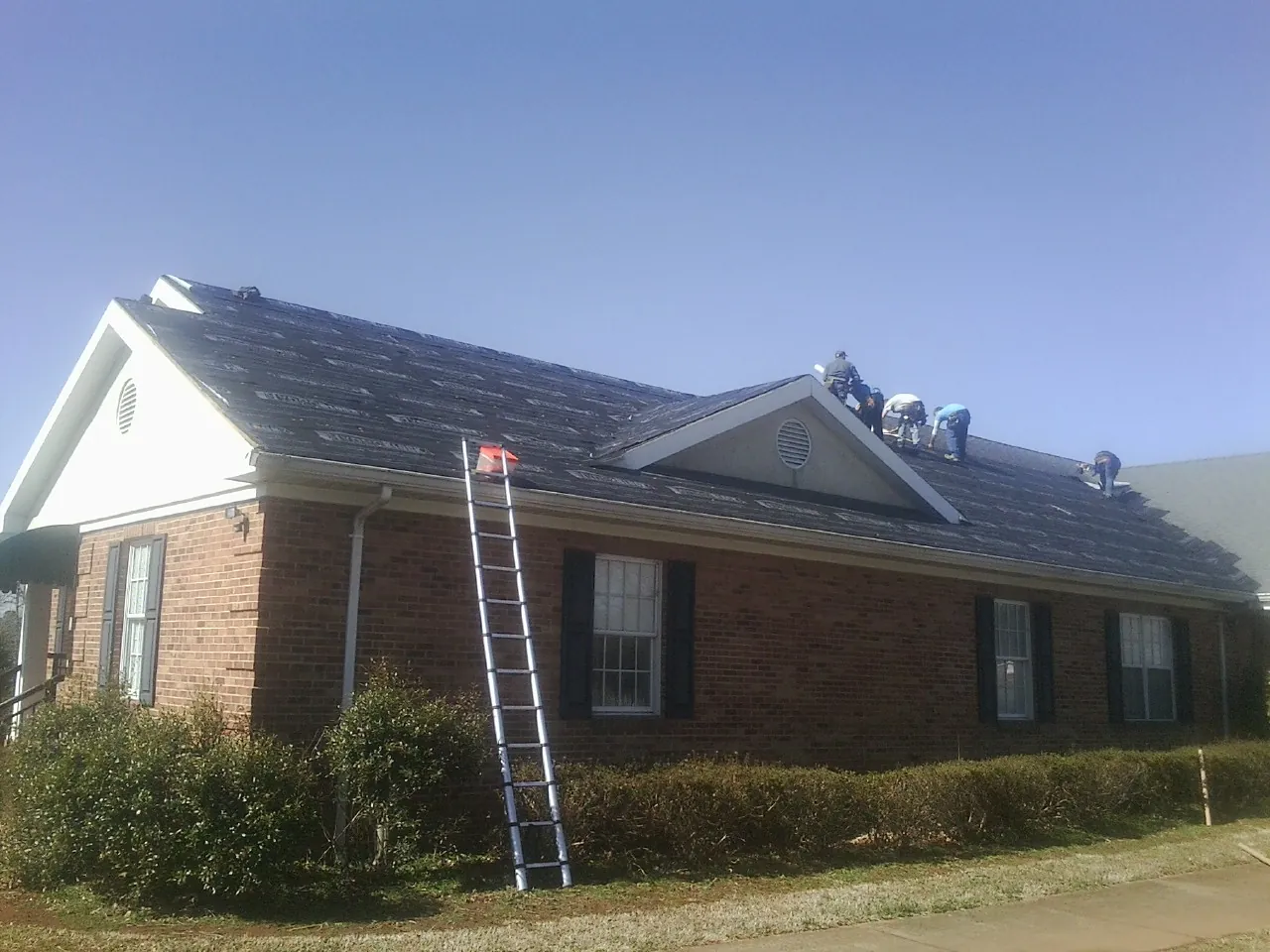 Crew tearing shingles off a brick ranch roof with a ladder set — Emergency Roof Repair work by our crew in Port Byron